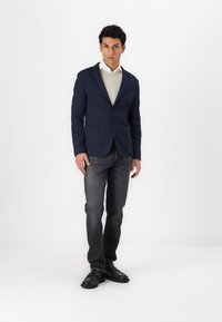 Navy blazer with two front pockets, worn over a light shirt, paired with dark jeans and black shoes, standing against a white background.