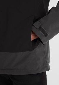 Black waterproof jacket with a grey bottom, featuring a side pocket and a Velcro strap at the wrist for adjustable fit. Smooth texture.
