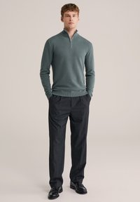 Žalias megztas megztinis su užtrauktuku prie kaklo, derinamas su formaliomis tamsiai pilkomis kelnėmis ir juodais batais, fone - neutrali spalva.