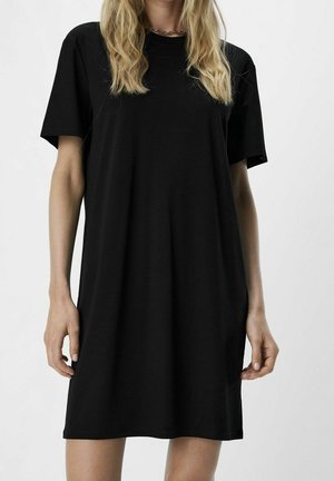 Mujer con un vestido tipo camiseta negro liso de manga corta, con cabello rubio ondulado y un collar de cadena plateada sobre un fondo liso.