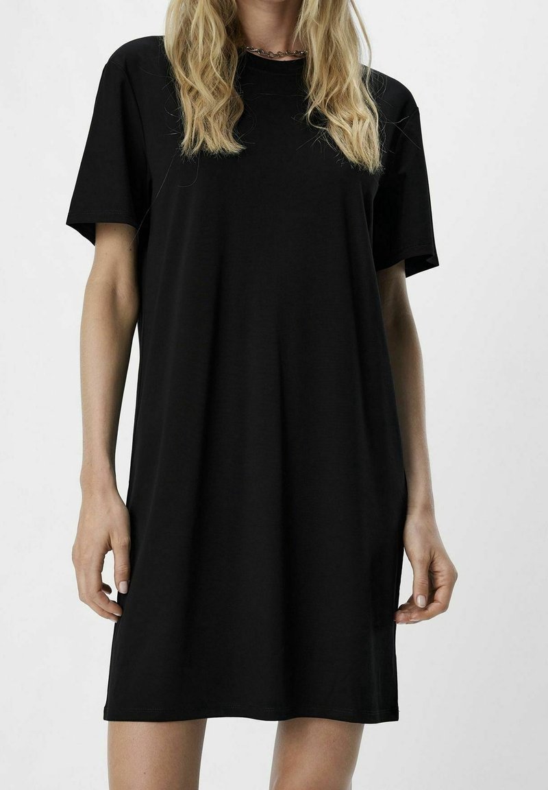 Femme portant une robe t-shirt noire unie à manches courtes, cheveux blonds ondulés et un collier chaîne argenté sur un fond uni.