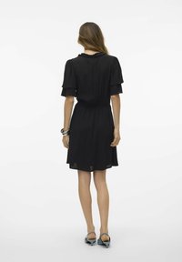 Svart klänning med korta, fjäderlätta ärmar, samlad midja och flödande kjol. Slät textur med volangkrage.