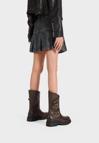 Stivaletti in pelle marrone con dettagli in fibbia e finitura testurizzata, indossati con una gonna a pieghe in denim. Lo sfondo è bianco uniforme.