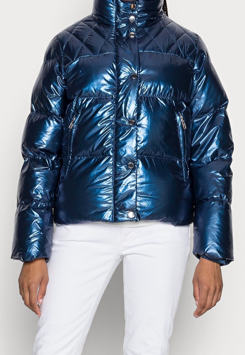 Personne portant une veste matelassée bleu marine brillante avec des boutons-pression et des poches zippées, associée à un pantalon blanc.
