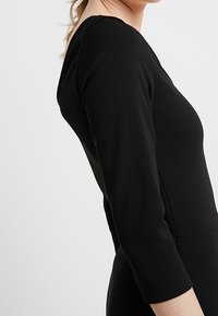 Svart långärmad klänning med en figursydd passform, rundad halsringning och mjuk textur. Sidovy visar ärmlängden och sömdetaljer.