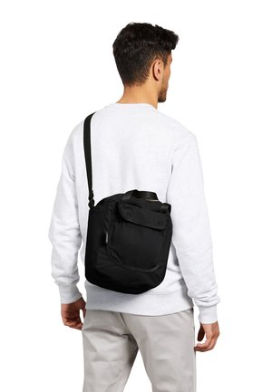 Homme portant un sweat-shirt blanc et un pantalon beige, portant un sac bandoulière noir avec une poche à rabat à l'avant sur son dos.
