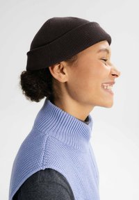 Bruine gebreide beanie, ronde vorm met ribbelstructuur, gecombineerd met een lichtblauw mouwloos sweater met een hoge kraag en zachte brei.