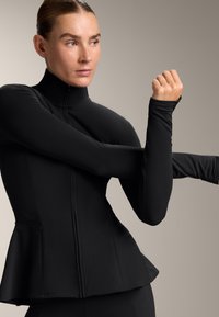 Black long-sleeve athletic jacket with a high collar, peplum design, and zip closure. Smooth fabric with fitted sleeves. Simple, modern silhouette.