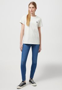 Camiseta blanca de algodón de manga corta con un pequeño logo azul marino en el pecho, acompañada de jeans ajustados azules y zapatillas negras.