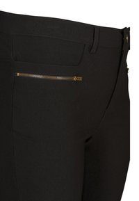 Schwarze Hose mit schmaler Passform, elastischem Material und einem goldenen Reißverschluss-Taschen-Detail auf der rechten Seite. Strukturierte Oberfläche.