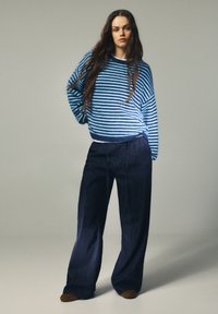 Modrý a biely pruhovaný pletený sveter s dlhými rukávmi v kombinácii s tmavomodrými nohavicami s širokými nohavicami. Oblečenie má voľný strih a minimálne zdobenie.