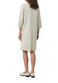 Helles graues knielanges Kleid mit 3/4-Ärmeln, geradem Schnitt und Rückennahtdetail; getragen mit hellen Schuhen, die einen niedrigen Absatz haben.