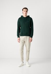 Felpa con cappuccio verde scuro realizzata in un tessuto morbido, con tasca frontale, cappuccio con cordino e logo quadrato. Abbinata a pantaloni jogger beige chiaro e sneakers bianche.