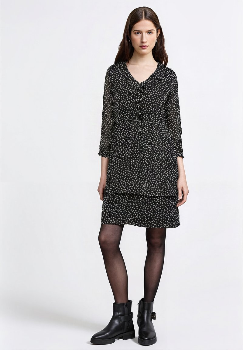 Jeune femme portant une robe noire à motifs floraux, des collants noirs transparents et des bottines noires, debout devant un fond blanc.