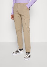 Beige cargo kalhoty mají několik kapes a rovný střih nohavic. Jsou nošeny s černo-bílými kostkovanými nazouvacími botami.