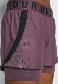 Lila Sportshorts mit schwarzen Akzenten, Mesh-Seitenteilen und einem breiten Bund mit dem Schriftzug "UNDER ARMOUR" in fettgedruckten schwarzen Buchstaben.