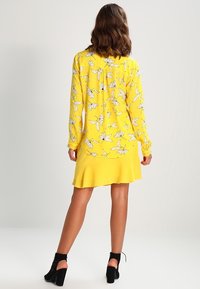 Gelbes Kleid mit einem weißen Blumenmuster, langen Ärmeln, einem ausgestellten Saum und einer weichen Stofftextur. Schwarze Knöchelstiefel vervollständigen den Look.