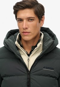 Ciemnozielona kurtka puchowa z kapturem, z zamkiem błyskawicznym i detalem z logo. Nosi się ją na kremowym swetrze dzianinowym oraz w kratkę koszuli.