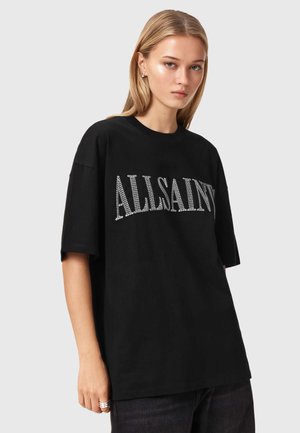 Jeune femme aux cheveux blonds raides portant un T-shirt noir surdimensionné avec le texte "ALLSAINTS" en grandes lettres blanches.