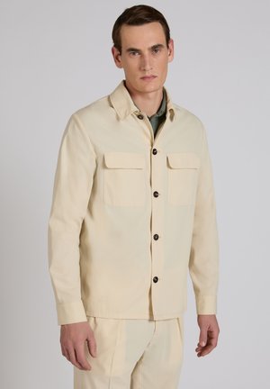 Mężczyzna ubrany w beżową kurtkę zapinaną na guziki z klapowymi kieszeniami, na jasnzie zielonej koszuli, stoi na jednolitym tle.