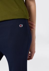 Granatowe legginsy z prążkowaną teksturą, z małym czerwono-białym logo na biodrze, zestawione z oliwkową górą.