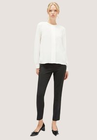 Weiße Bluse mit langen Ärmeln, runder Halslinie und vertikalen Faltendetails, kombiniert mit schmalen schwarzen Hosen und flachen Schuhen mit schwarzer Schleifenverzierung.