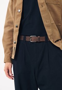 Cinturón de cuero marrón con hebilla plateada, usado sobre pantalones oscuros y una camisa negra, complementado con una chaqueta abotonada de color marrón claro.