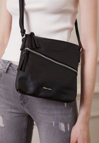 Černá texturovaná kožená crossbody taška s diagonálním zipem, stříbrnými detaily a dvěma třásněmi. Kompaktní obdélníkový tvar.