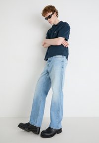 Jeans azzurri chiari, comodi e dal finish sbiadito, abbinati a una camicia a maniche corte blu scuro con bottoni. Zoccoli neri e occhiali da sole completano il look.