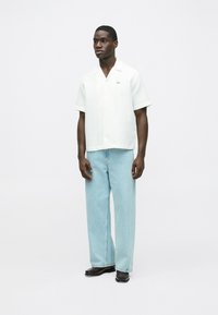 Biela textúrovaná košeľa s krátkym rukávom a golierom, kombinovaná s ľahkými modrými džínsami s širokými nohavicami a čiernou obuvou. Na košeli je nenápadný detail v podobe loga.