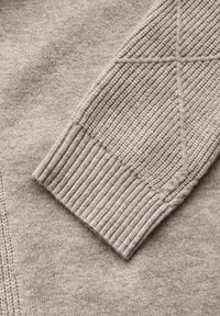 Beige stickad tröja detalj, med ribbad manschett och diamantskärningstextur på ärmen. Mjuk väv med en slät yta.