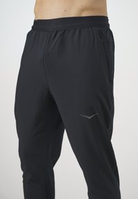 Pantaloni de trening negri, cu talie elastică, croi conic, buzunar lateral și un logo discret în argintiu pe piciorul stâng.