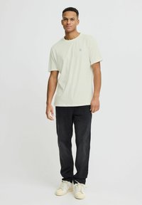 Camiseta de algodón verde claro con cuello redondo y mangas cortas, combinada con jeans negros y zapatillas blancas. Pequeño logo azul en el pecho.