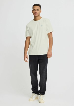 Camiseta de algodón verde claro con cuello redondo y mangas cortas, combinada con jeans negros y zapatillas blancas. Pequeño logo azul en el pecho.