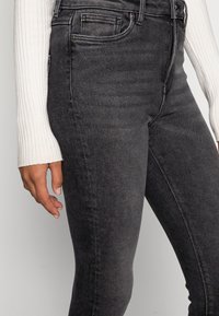 Jeans ajustados gris oscuro con una textura suave, diseño de cinco bolsillos y cierre de cremallera. Presenta un ajuste ceñido en el tobillo.
