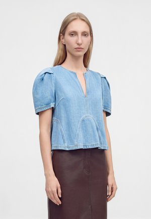 Mujer con pelo rubio liso lleva una camiseta de mezclilla azul claro de manga corta con hombros abullonados y una falda marrón sobre un fondo liso.