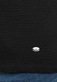Zwarte gebreide trui met horizontale ribbels, voorzien van een klein zilveren logo-plaatje aan de onderkant. De stof lijkt zacht met een gebreide afwerking.