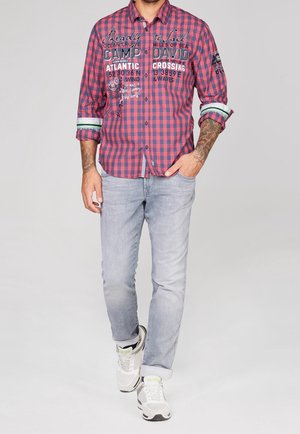 Camisa de manga larga con cuadros rojos y azules, con texto y gráficos, combinada con jeans gris claro y zapatillas blancas.