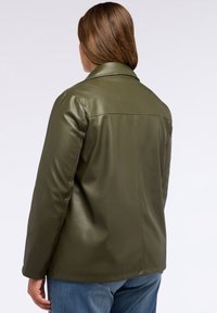 Person som vänder ryggen mot kameran, if örd en olivgrön läderjacka och blå jeans mot en helt vit bakgrund.