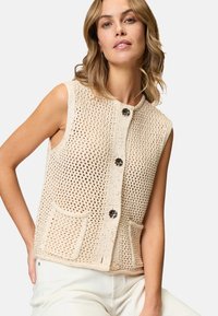 Strikket ærmeløs vest i beige, med et åbent vævdesign, to forlommer og knaplukning med dekorative hardwaredetaljer.