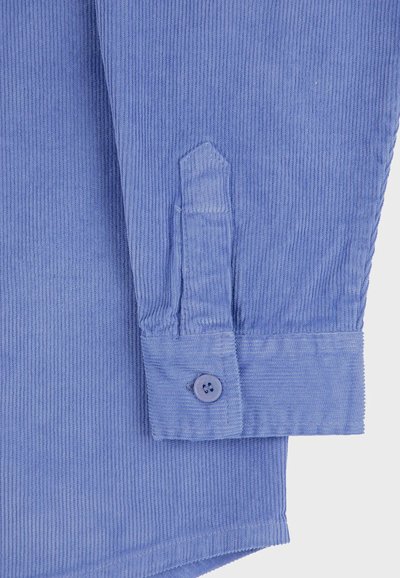 Tessuto di camicia in corduroy blu con texture a righe verticali, caratterizzato da un polsino con bottoni e un taglio angolare all'orlo delle maniche.