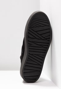 Černá semišová podrážka obuvi s texturovanou gumovou základnou a diagonálními drážkami, nakloněná proti bílému pozadí.