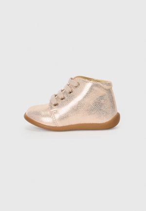 Chaussure bébé métallique rose brillante avec semelle marron et lacets beiges, vue de côté sur un fond clair uni.