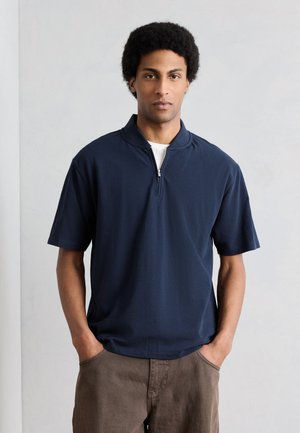 Giovane uomo con capelli corti e ricci, che indossa una maglietta blu navy a maniche corte con cerniera e pantaloni marroni, in piedi con le mani in tasca.