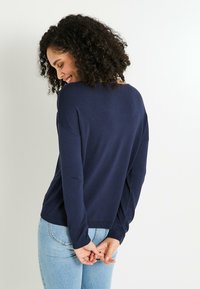 Joven mujer con cabello rizado, vistiendo un suéter azul marino y jeans de un azul claro, sonriendo y mirando hacia abajo con las manos entrelazadas detrás de su espalda.