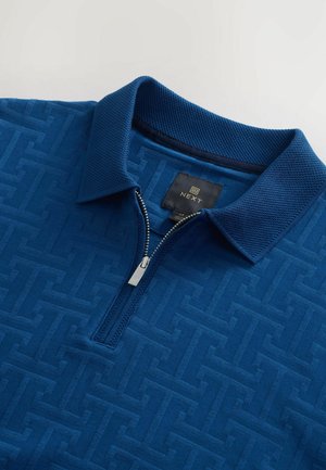 Blue zip-up polo shirt featuring textured fabric with a geometric pattern, a collared neck, and a silver zipper. Brand label visible.