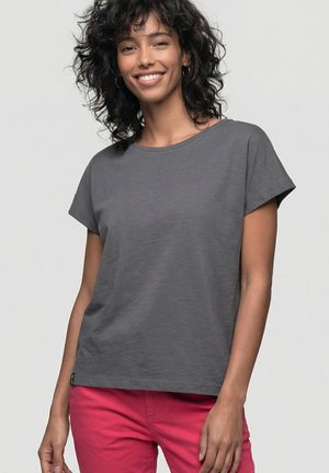 Lächelnde Frau mit lockigem Haar, die ein schlichtes graues T-Shirt mit kurzen Ärmeln und leuchtend pinke Hosen trägt, vor einem hellgrauen Hintergrund stehend.