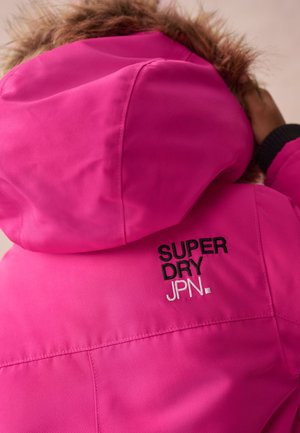 Roza vodoodporna jakna s kapuco obrobljeno z krznom. Na zadnji strani ima izvezen napis "SUPER DRY JPN." Teksturna tkanina z gladkim zaključkom.