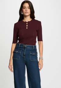 Eine Frau mit langen, dunklen Haaren trägt ein kastanienbraunes, kurzärmliges Strickoberteil und hochgeschnittene blaue Jeans mit Klapptaschen vorne.