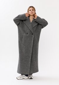 Lungo cappotto grigio oversize realizzato in tessuto testurizzato, con un ampio colletto, chiusura doppiopetto e grandi tasche laterali.